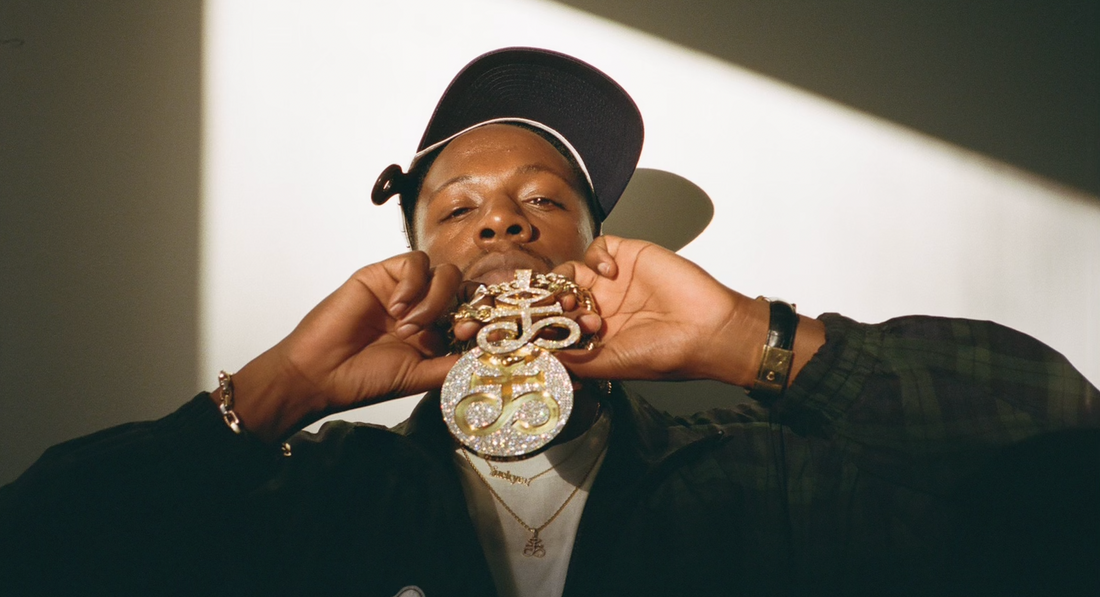 Brooklyn rapper Joey Bada$$ holding a custom white gold diamond nameplate necklace designed for his sister, featuring pavé diamond lettering, engraved back, and tennis chain — crafted in NYC’s Diamond District as a luxury family gift.